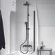 Duschsystem in Schwarz mit feststehendem Kopf- und Handbrause an einer gläsernen Duschwand. Neben der Dusche steht ein Beistelltisch mit Accessoires. Ideal Standard.